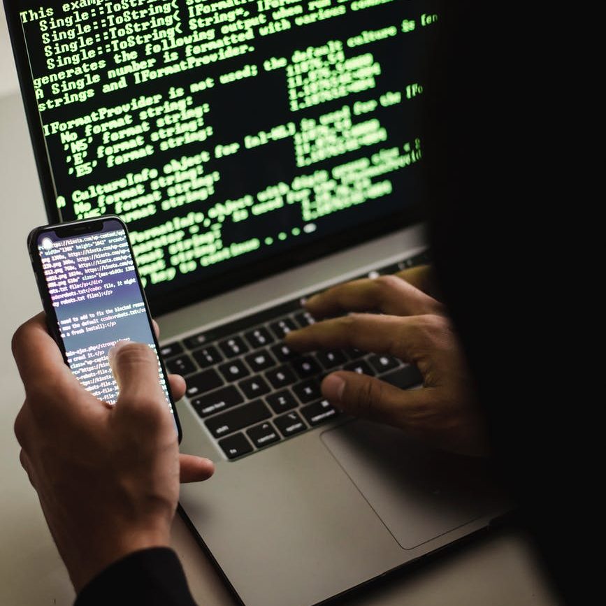 unrecognizable hacker with smartphone typing on laptop at desk